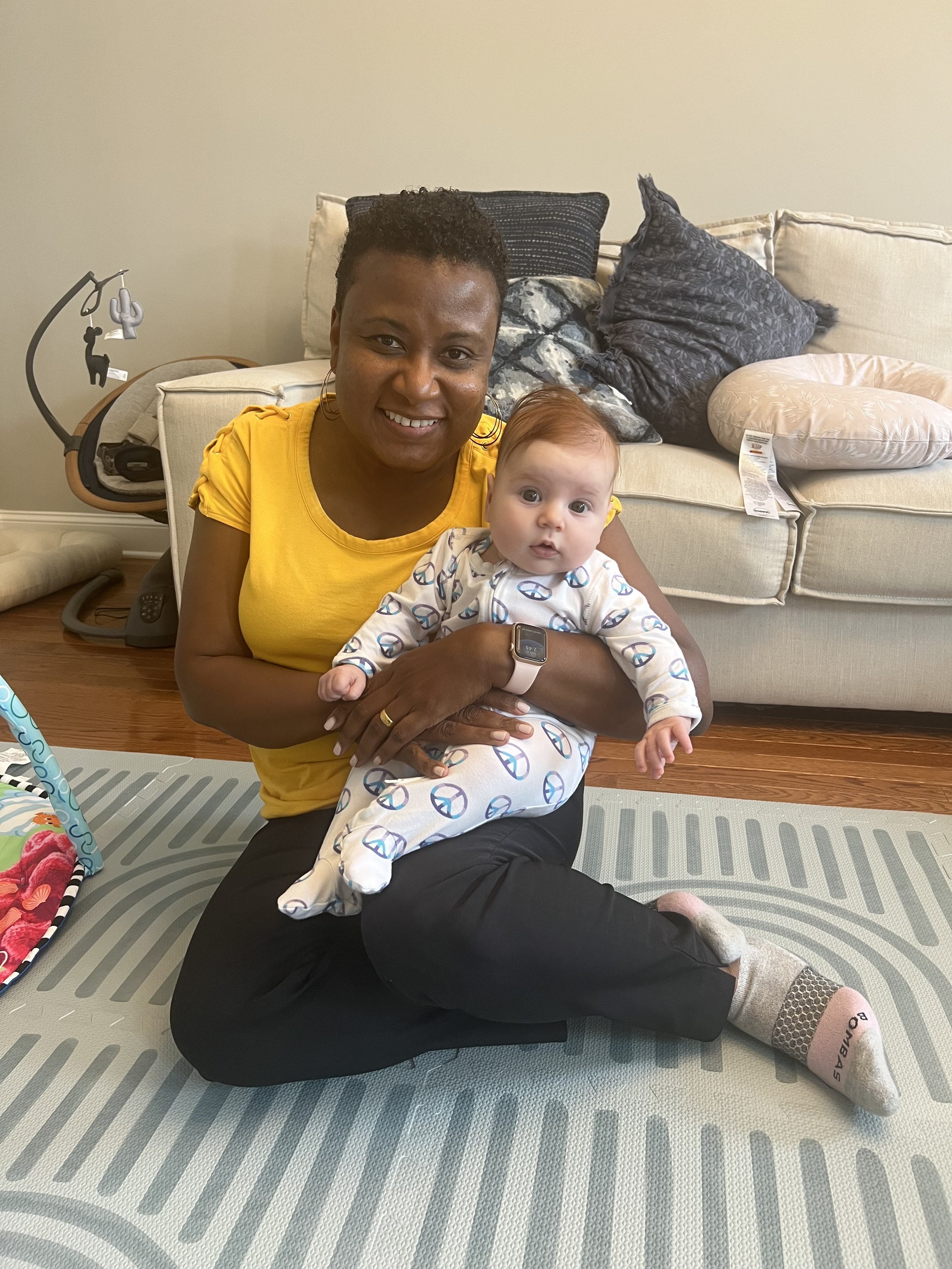 Ria Savoy, birth and postpartum doula holding a smiling baby in front of a couch at client home in Philadelphia.