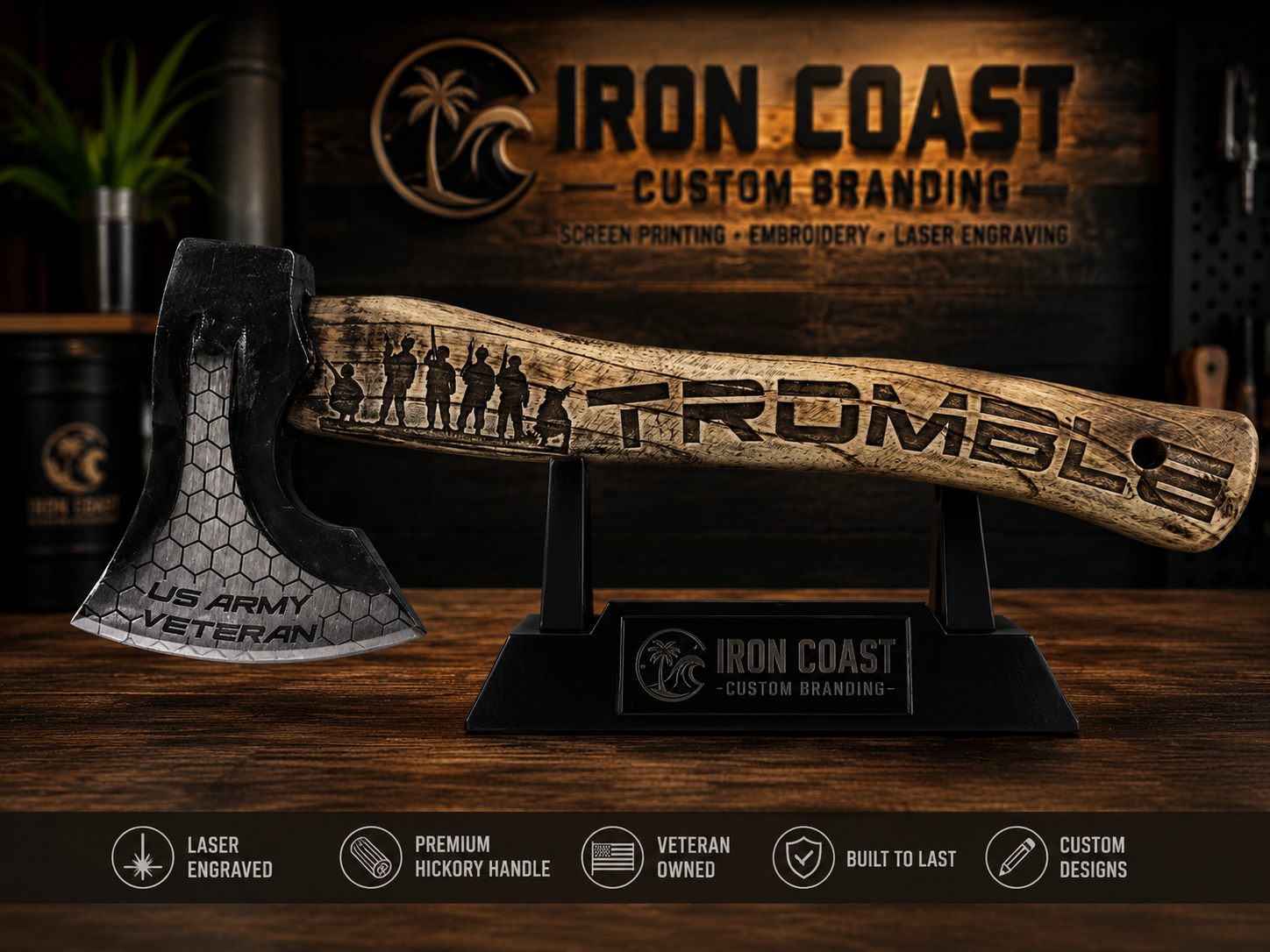 An axe with a wooden handle engraved with silhouettes of soldiers and the word 'TRUMP' on it, displayed on a black stand with a logo for Iron Coast Custom Branding in the background.