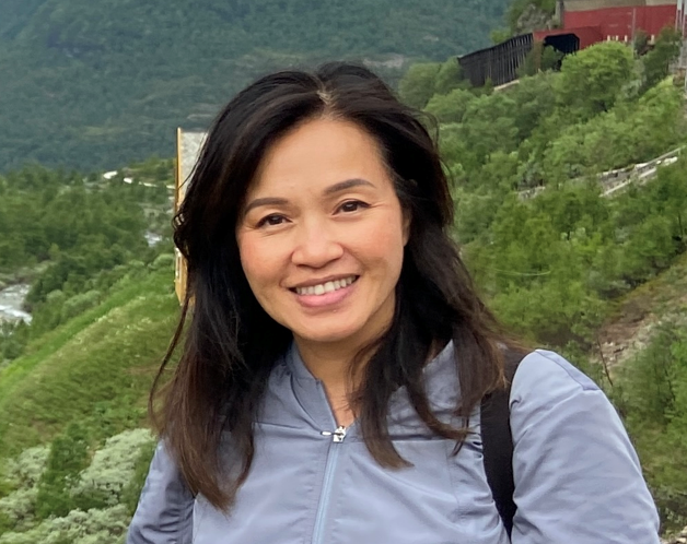 Smiling woman with shoulder-length dark hair wearing a light gray jacket outdoors with green hills and trees in the background.