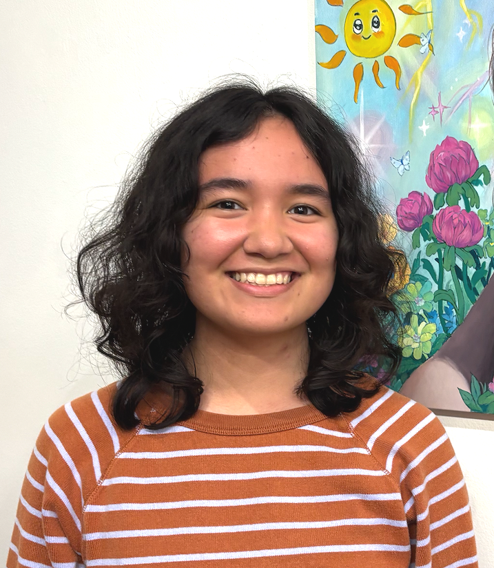 A young woman with curly black hair and a big smile standing in front of colorful artwork featuring a smiling sun and pink flowers.