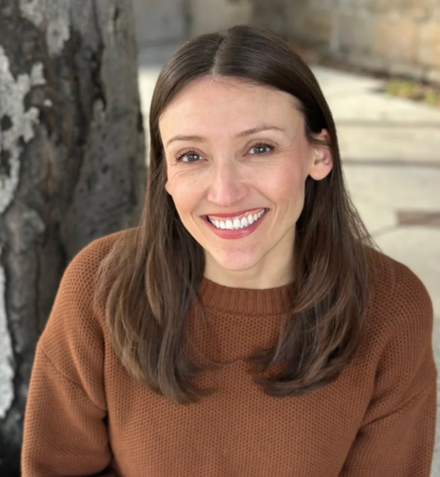 Woman with long brown hair and a brown sweater smiling outdoors near a tree.