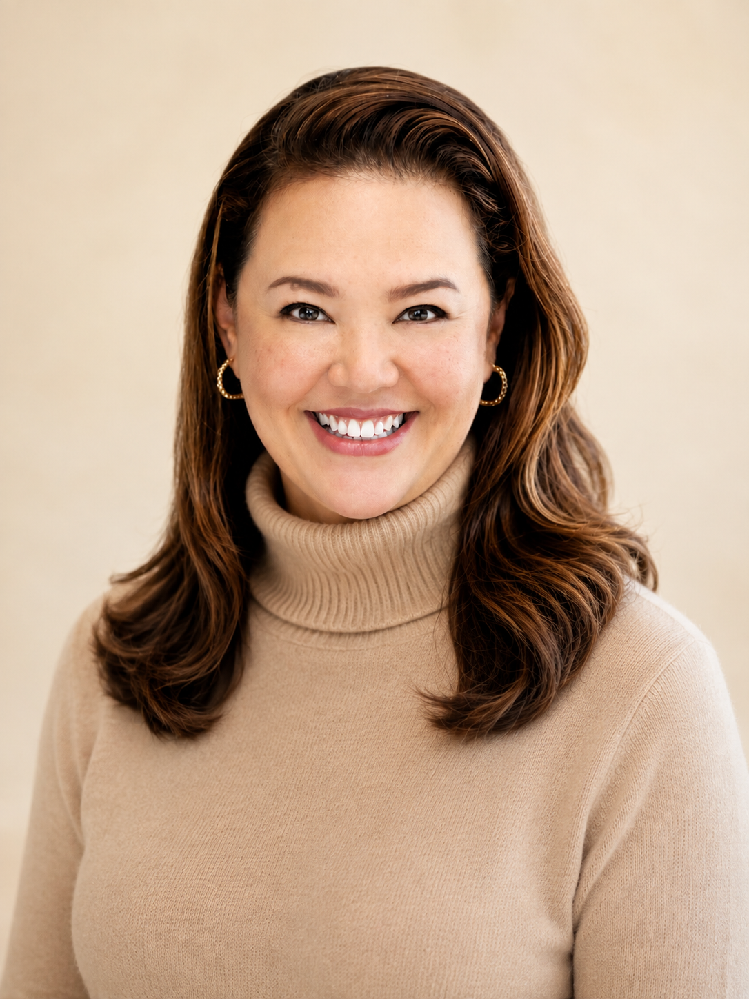 A woman with brown, wavy hair wearing a beige turtleneck sweater and gold hoop earrings, smiling at the camera.