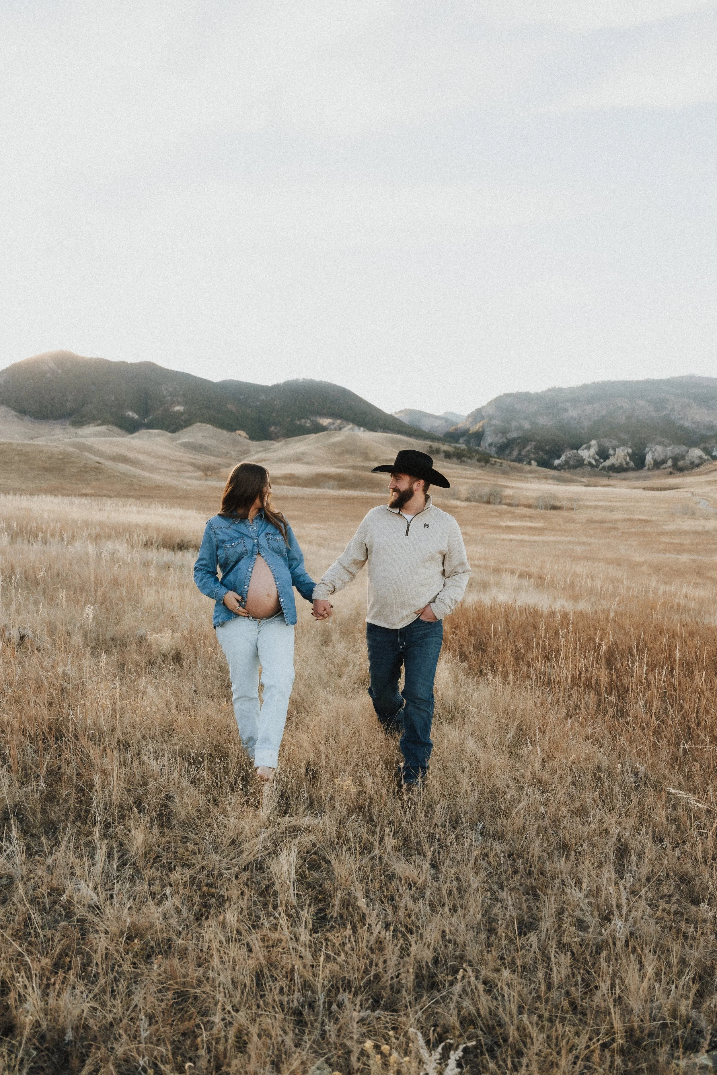 A pregnant woman and a man holding hands, walking through a grassy field with mountains in the background.