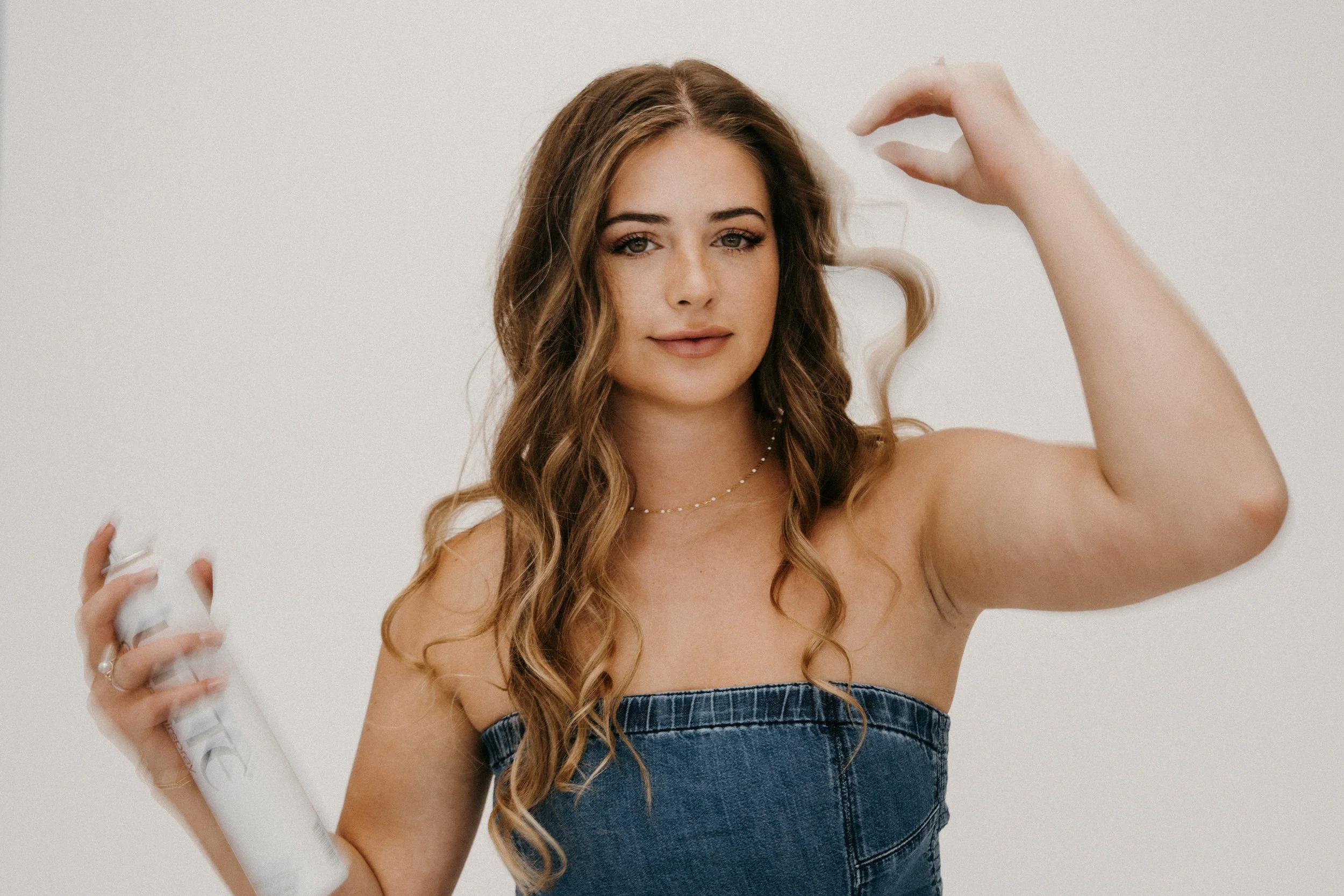 A woman with long, wavy brown hair wearing a strapless denim top, holding a can of dry shampoo in her right hand, and spraying it onto her hair.