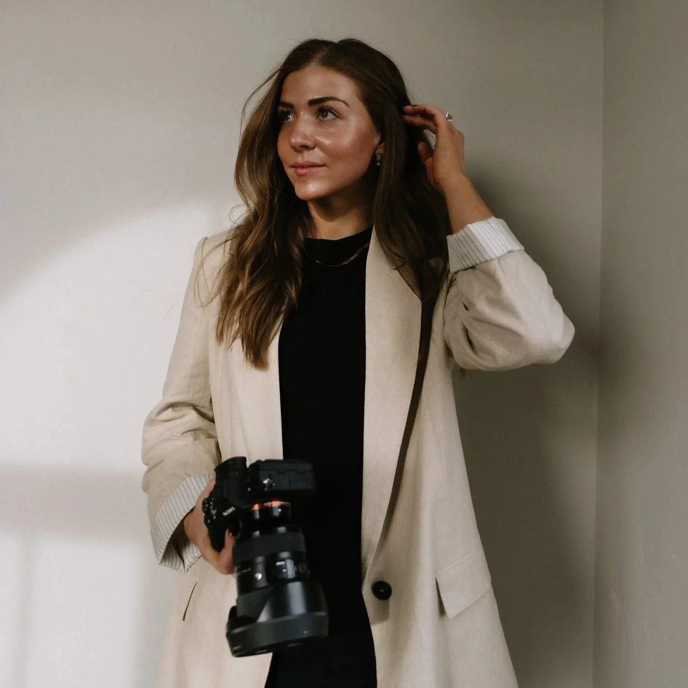 A woman with long brown hair, wearing a beige blazer over a black top, holding a camera, and touching her hair, standing against a plain wall.
