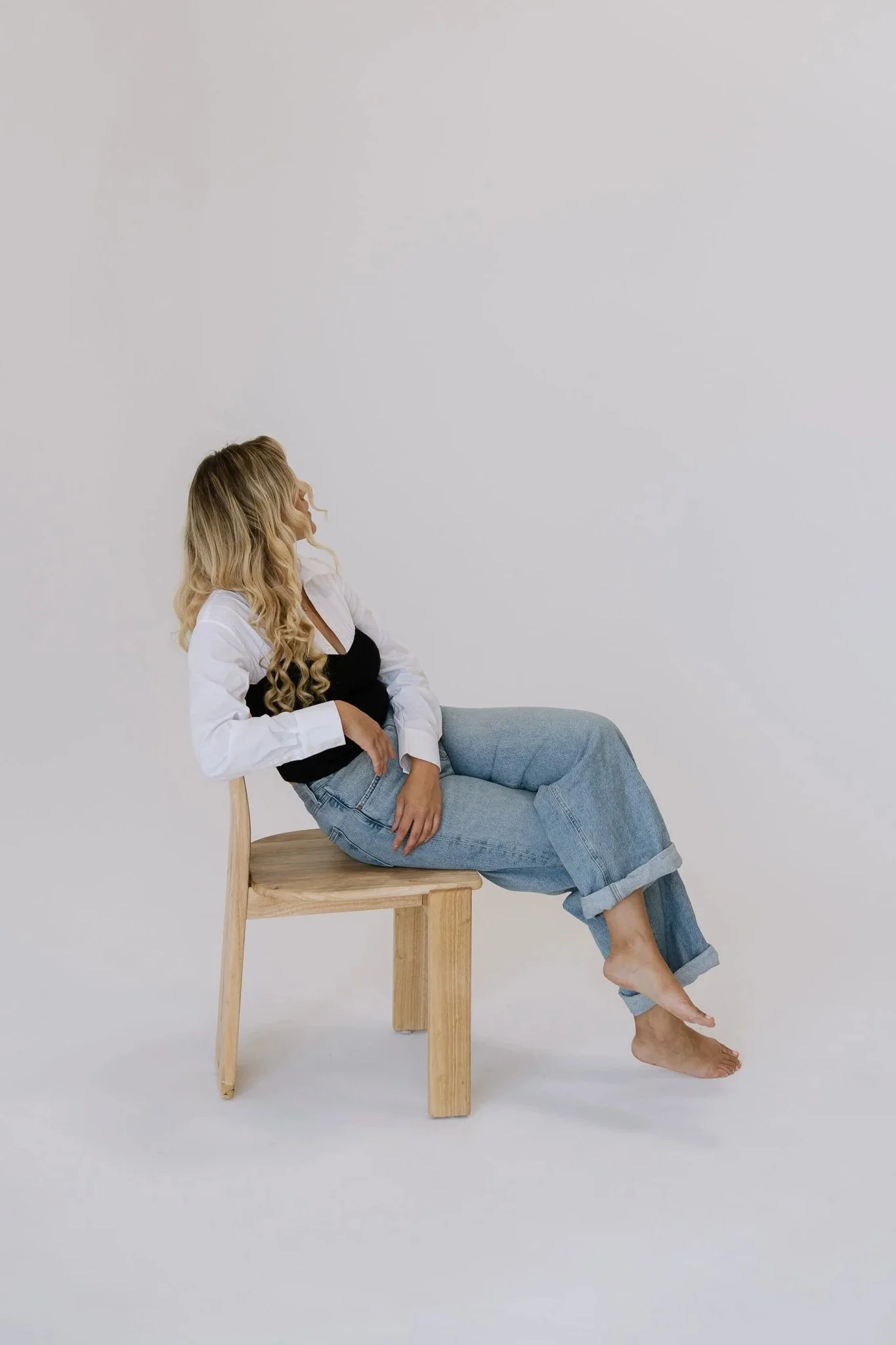 A woman with long wavy blonde hair sitting on a wooden chair with her legs crossed, wearing a white shirt, black top, and rolled-up jeans, against a plain light background.