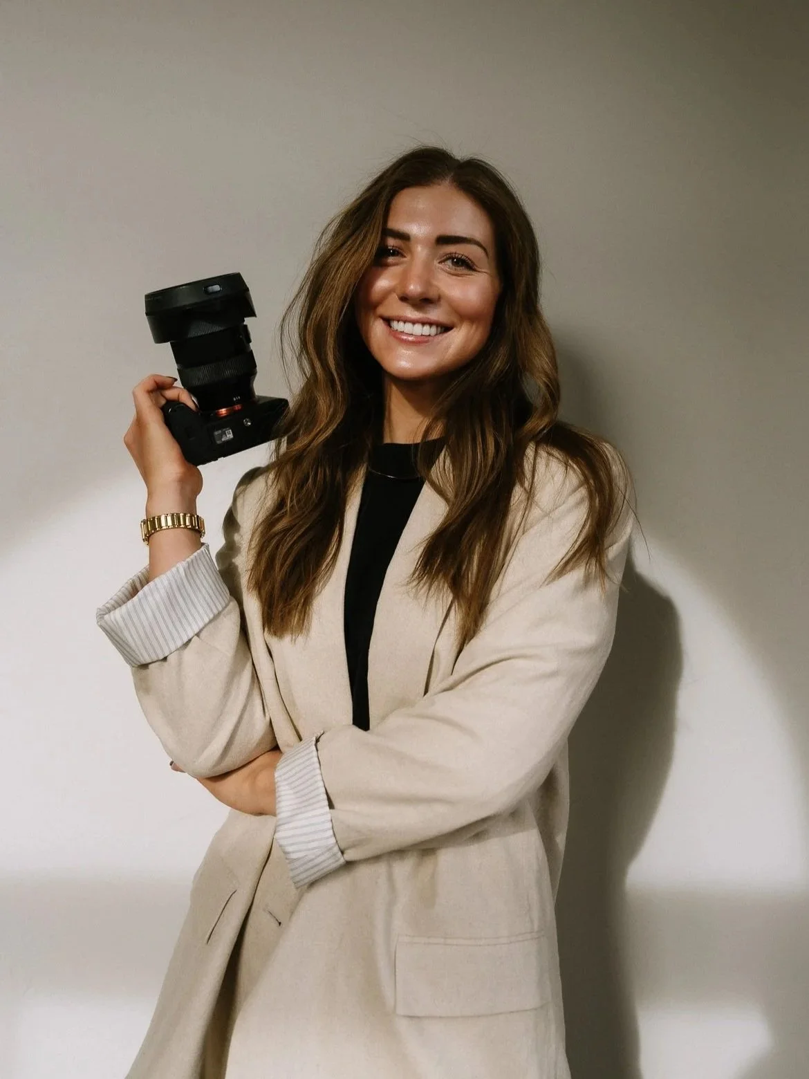 A woman with long brown hair and a beige blazer holding a camera with a large lens, smiling at the camera against a plain background.