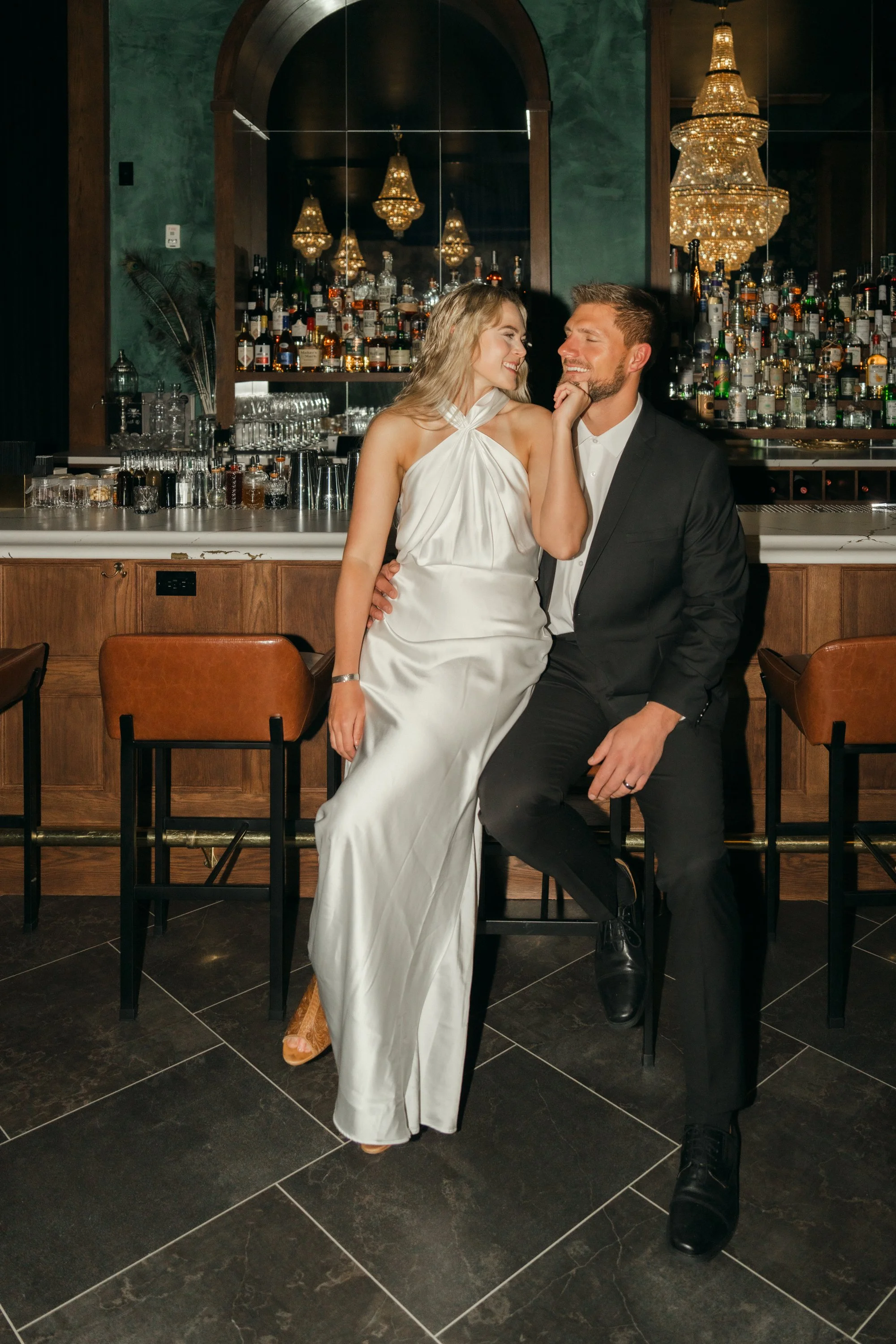 A couple in wedding attire sitting at a bar, the woman in a white wedding dress and the man in a black suit, smiling and looking at each other.