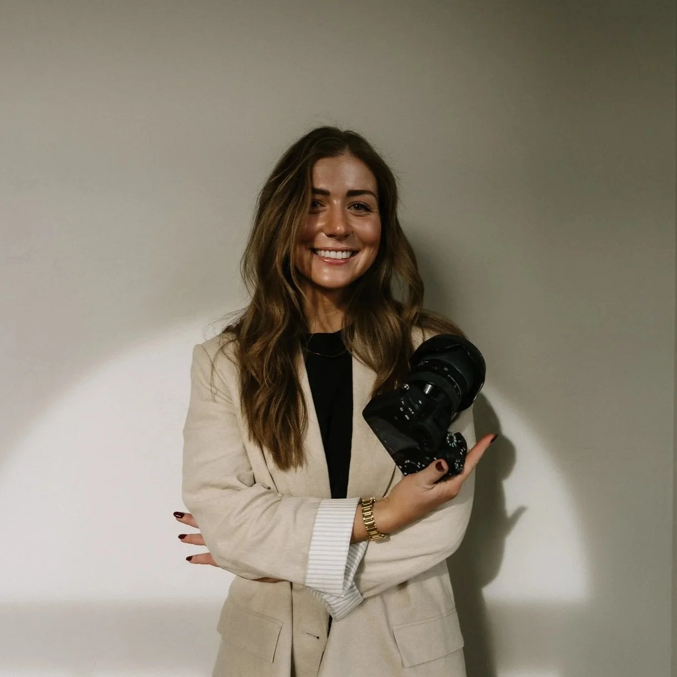 A woman with long wavy brown hair, smiling, holding a professional camera in her right hand, standing against a plain light-colored wall