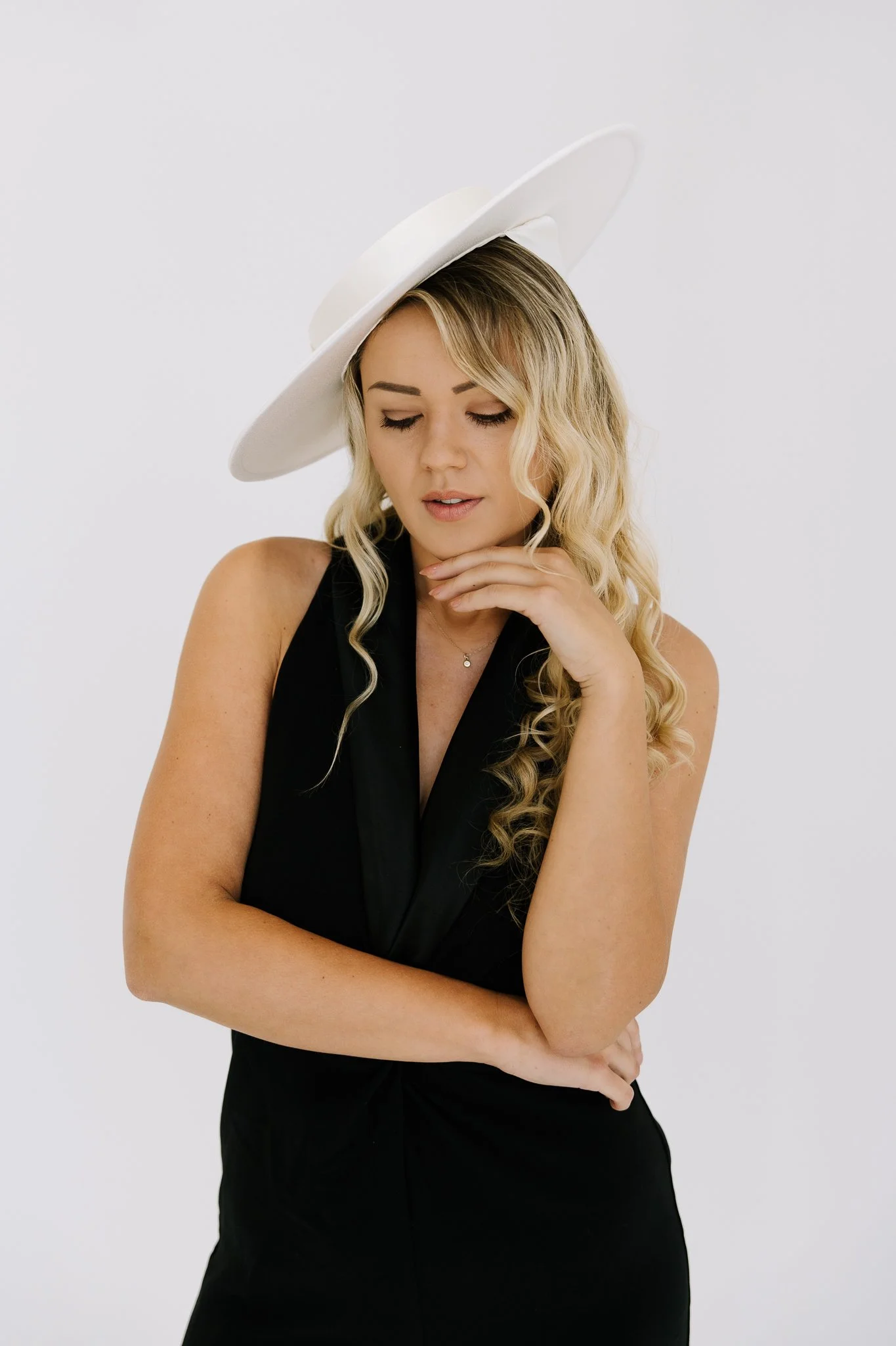 Woman with long, wavy blonde hair wearing a white wide-brimmed hat and black sleeveless dress against a plain white background.