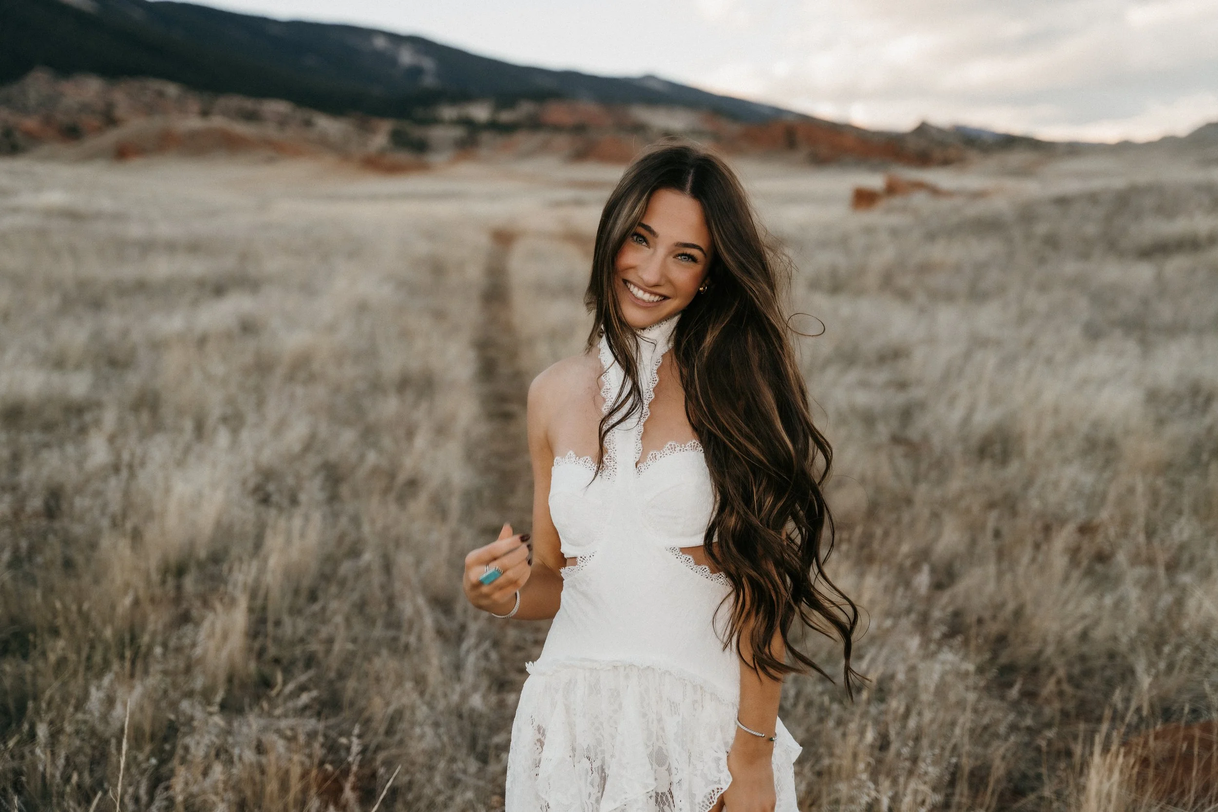 A woman with long wavy brown hair smiling and wearing a white lace dress standing in a field of dry grass with mountains in the background.