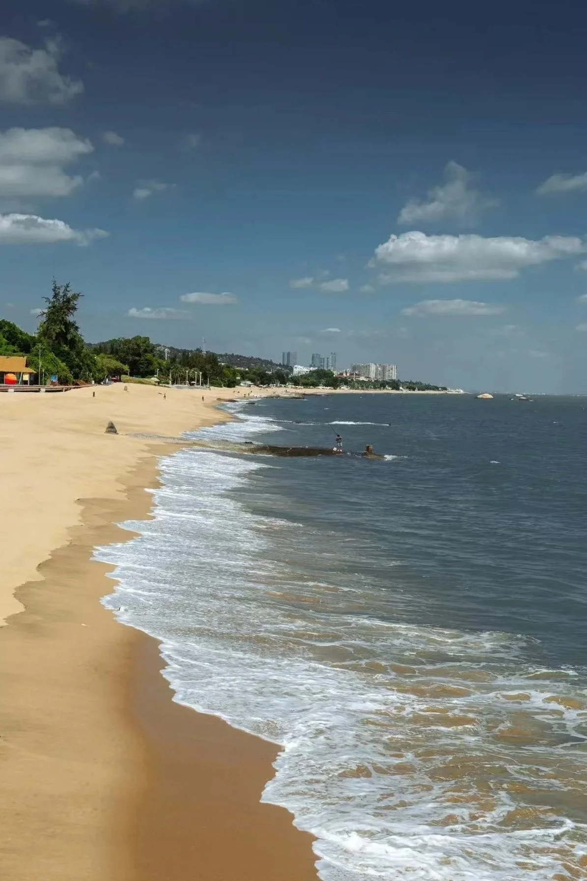 Beach with golden sand, blue water, and some people fishing along the shoreline. City buildings are visible in the distance under a partly cloudy sky.