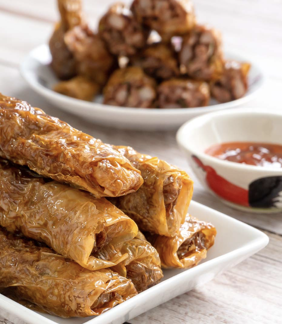 Plate of freshly rolled, cooked spring rolls with a side of dipping sauce, and a bowl of meatballs in the background.