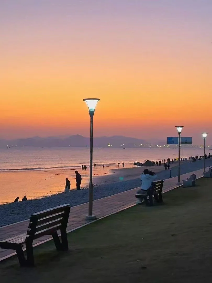 Beachside promenade during sunset with lampposts, benches, and people strolling along the shore and sitting on benches, calm water with distant mountains and city lights