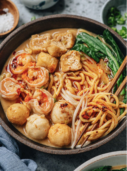 A bowl of ramen with shrimp, fish balls, tofu, leafy greens, and noodles in broth, with chopsticks and bowls of rice and green onions visible in the background.