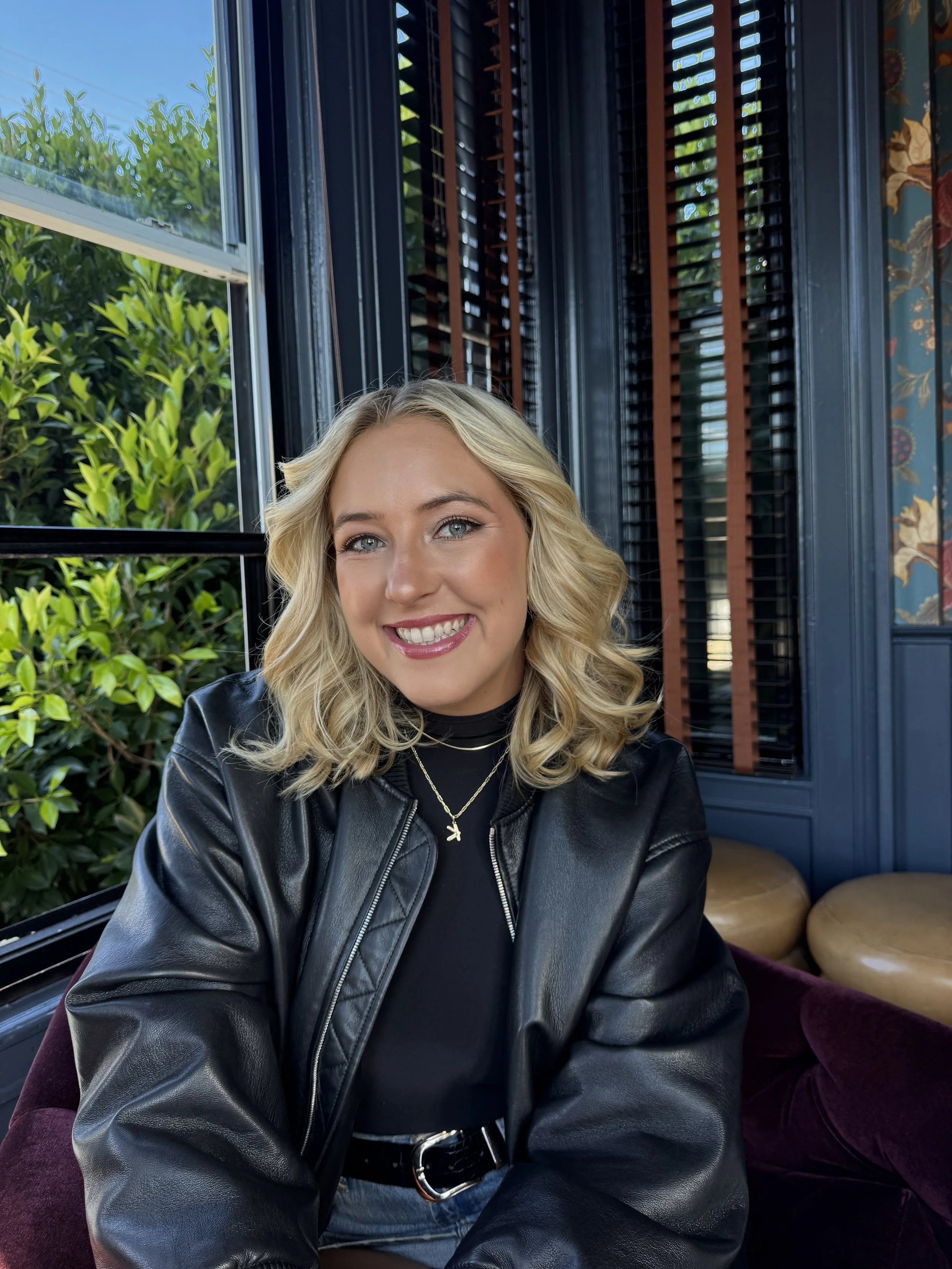 A woman with blonde curly hair and blue eyes smiling, wearing a black leather jacket and layered necklaces, sitting indoors near a window with greenery outside.