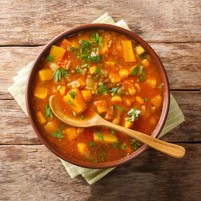 Bowl of vegetable soup with diced carrots, squash, and herbs, on a wooden table with a wooden spoon.