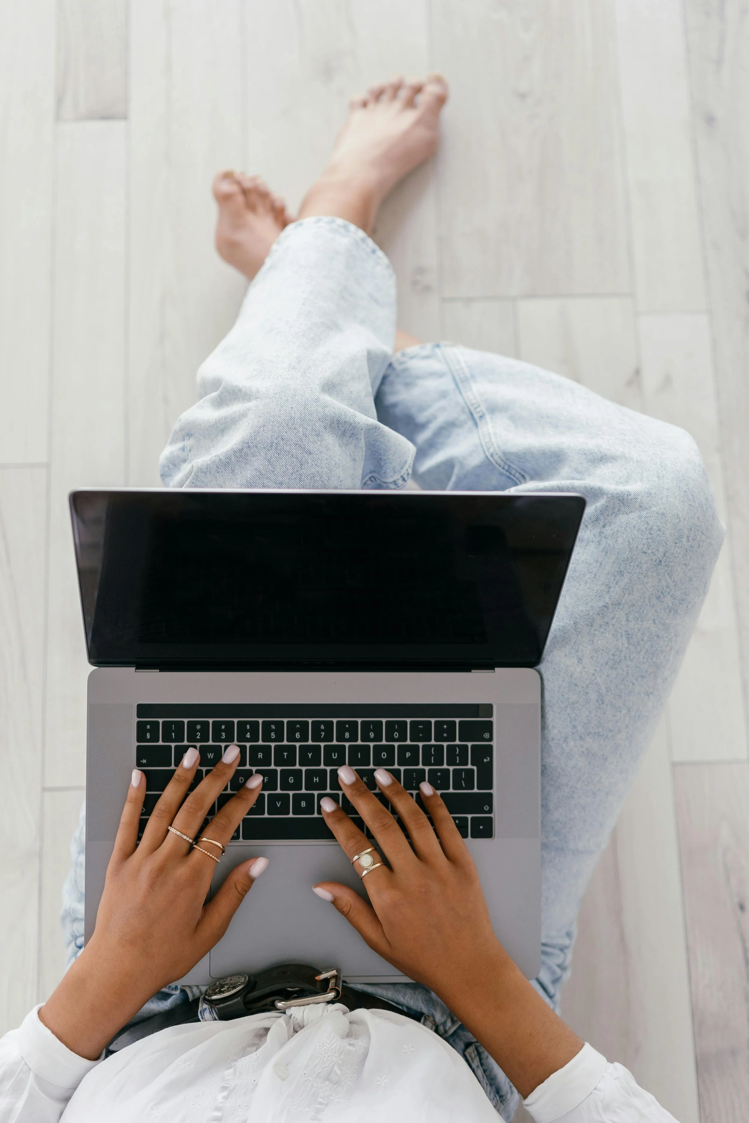 From an overhead view, a person with painted nails and rings on their fingers is typing on a laptop placed on their lap, while another person with bare feet and denim jeans sits on the floor nearby, with their legs crossed and hands resting on their knees, in a light-colored room.