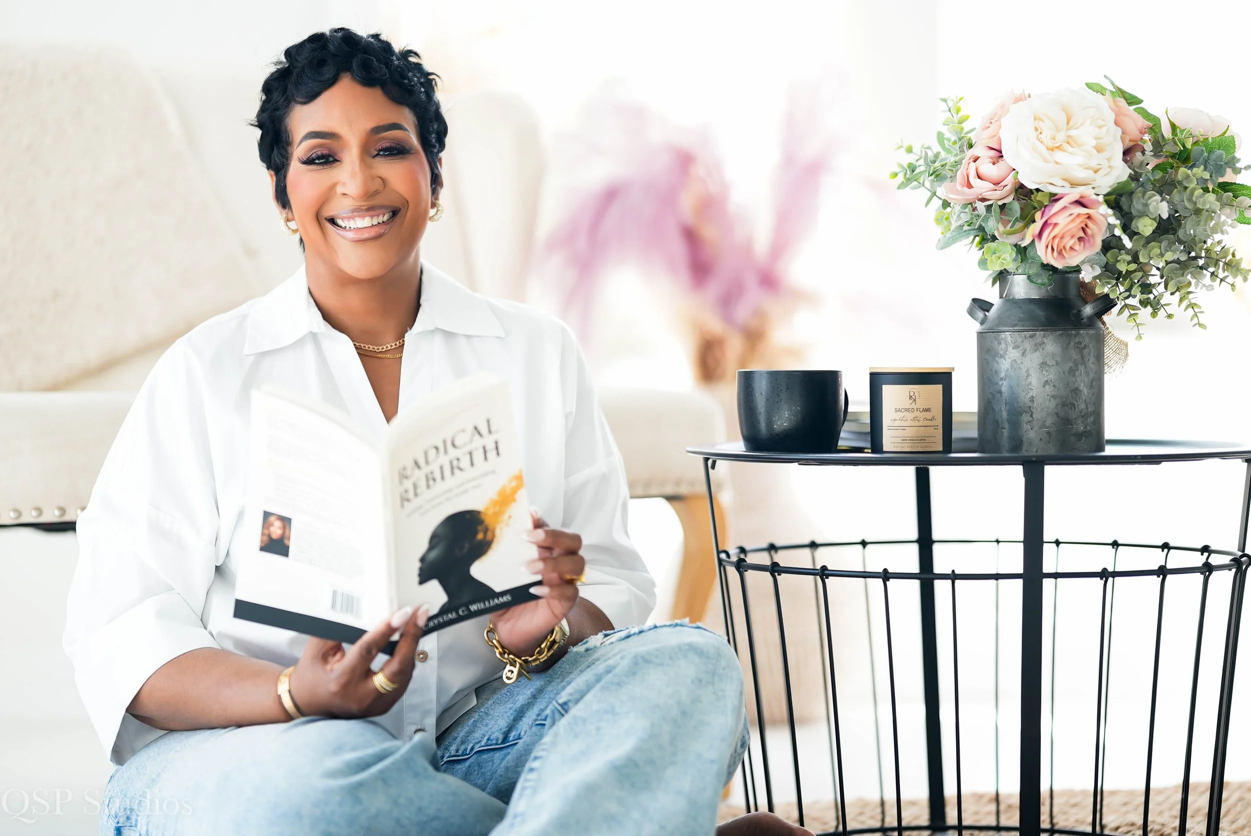 Woman sitting on a chair, smiling and holding a book titled 'Radical Rebirth', with an open book in her lap, wearing a white shirt and gold jewelry, next to a table with a large vase of pink and white roses and a black scented candle, in a bright room.