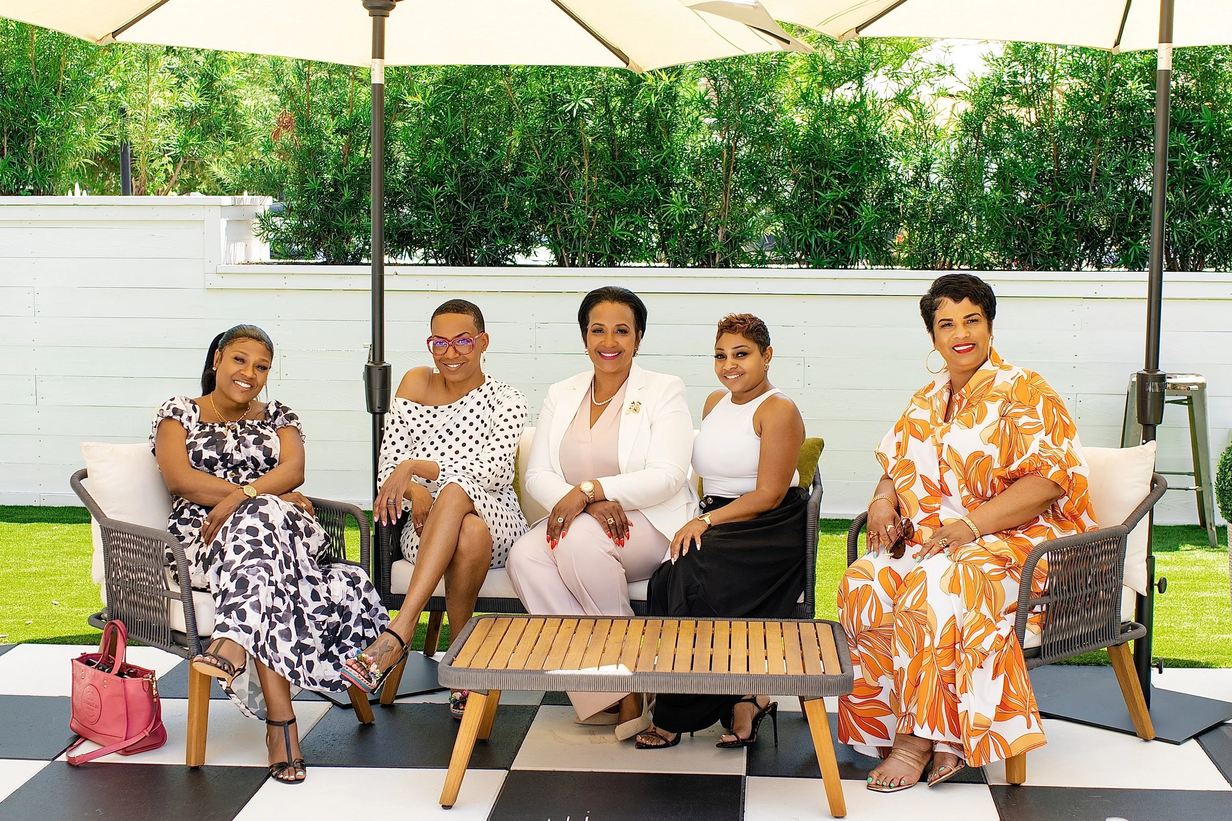 Six women sitting outdoors under large umbrellas on a patio with a white fence and green foliage in the background. One woman is wearing a white blazer, others are dressed in patterned or colorful outfits, and they are smiling at the camera.