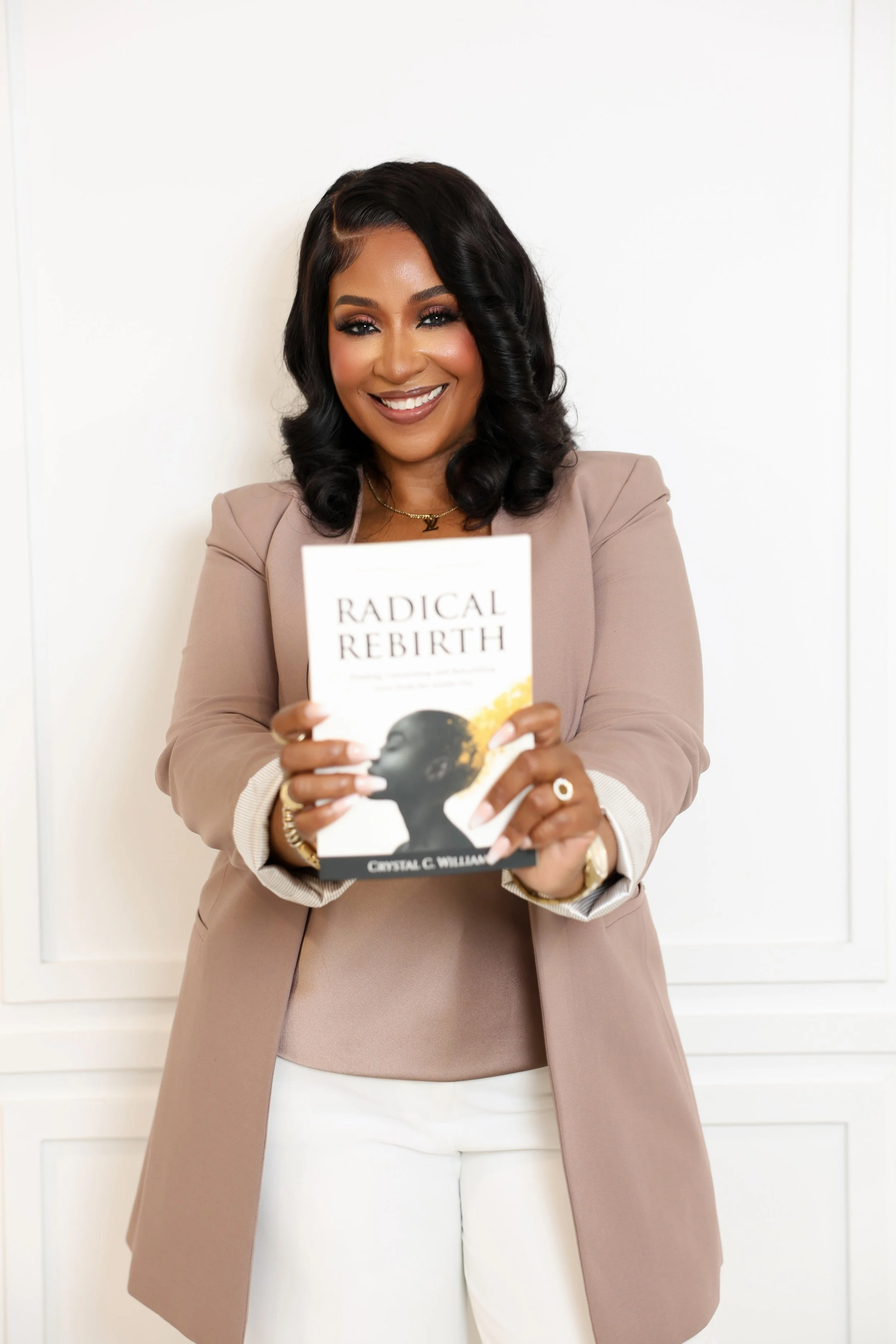 Woman smiling, holding a book titled 'Radical Rebirth' by Crystal C. Williams, in front of a plain white wall.