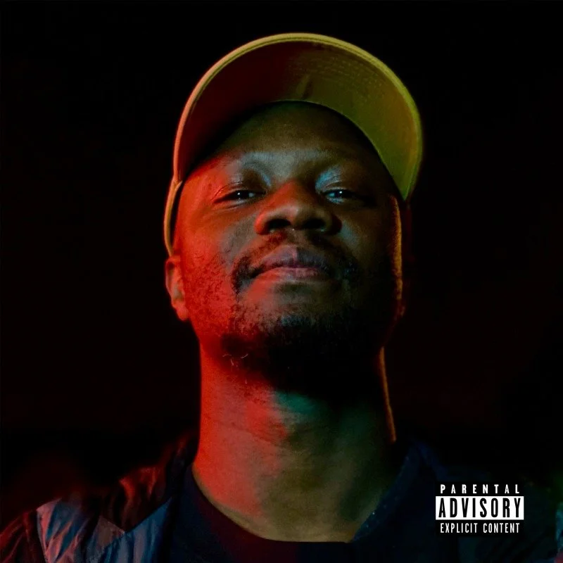 Album cover for "Rigganish" A man wearing a white cap, looking directly at the camera, illuminated with red and green lighting against a dark background.