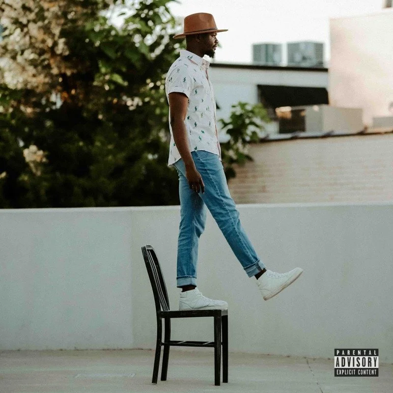 A man walking on a chair on a rooftop with trees and buildings in the background, wearing a hat, white patterned shirt, blue jeans, and white sneakers.