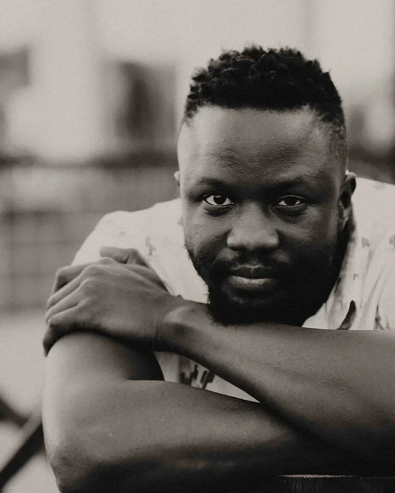 A black-and-white portrait of a man with short, curly hair and a beard, resting his head on his crossed arms and looking directly at the camera.