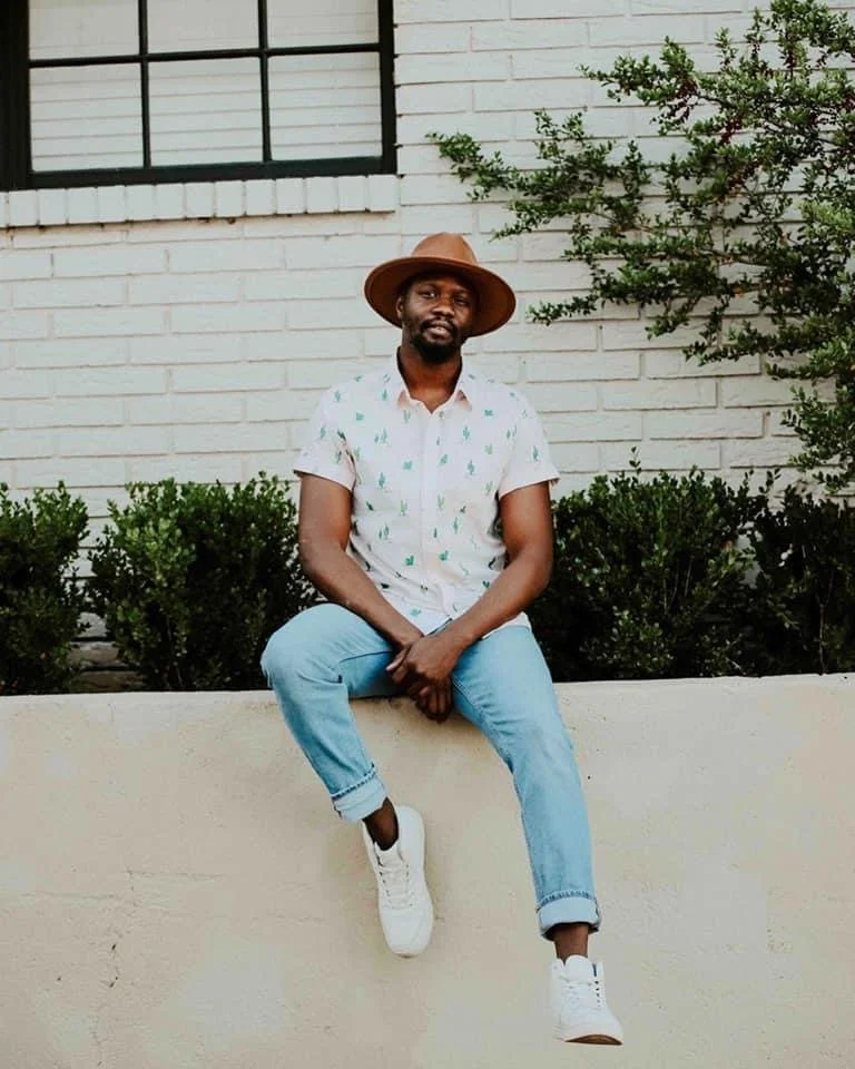 A man sitting on a white wall outside a building with a large window, wearing a beige hat, white short-sleeve shirt with green patterns, light blue jeans, and white sneakers.