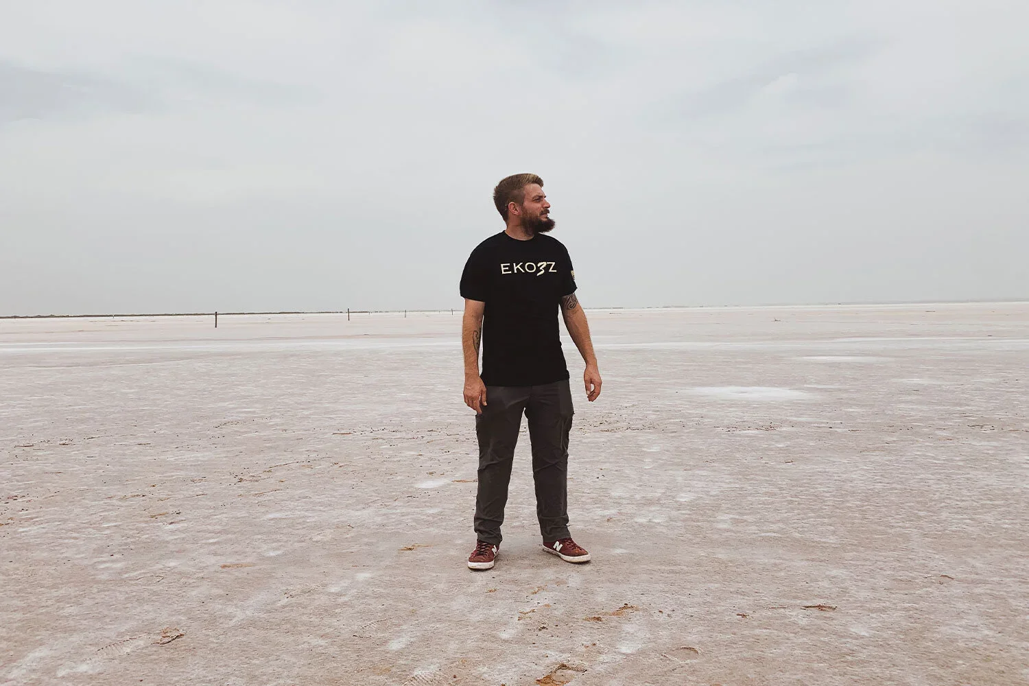 Man standing in a vast, flat salt desert with an overcast sky, wearing a black T-shirt, dark pants, and red sneakers.