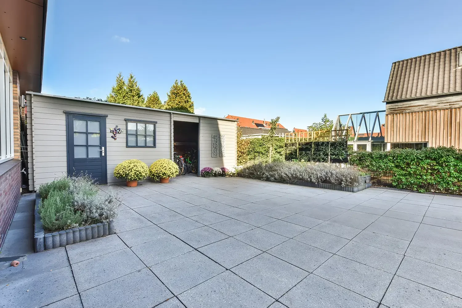 Clean backyard with large gray tiled patio, shed with storage of bicycles, potted plants, and well-maintained shrubs and garden beds, under a clear blue sky.