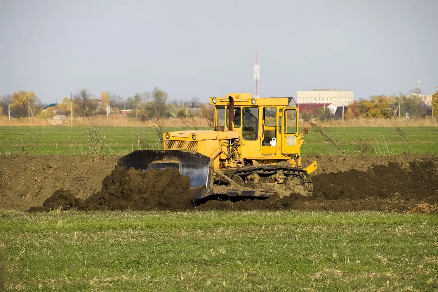A yellow bulldozer pushing soil in a field with a background of trees and buildings.