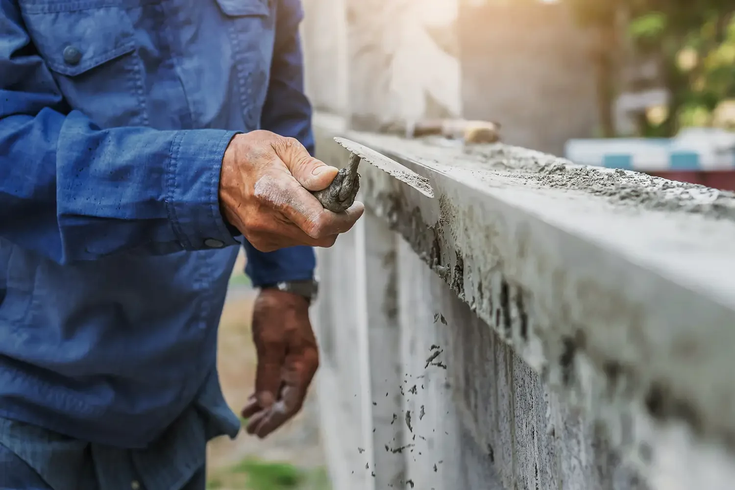 Person in blue denim shirt using a trowel to apply cement or plaster to a wall, with a focused hand and part of their arm visible, outdoors with sunlight and blurred background.