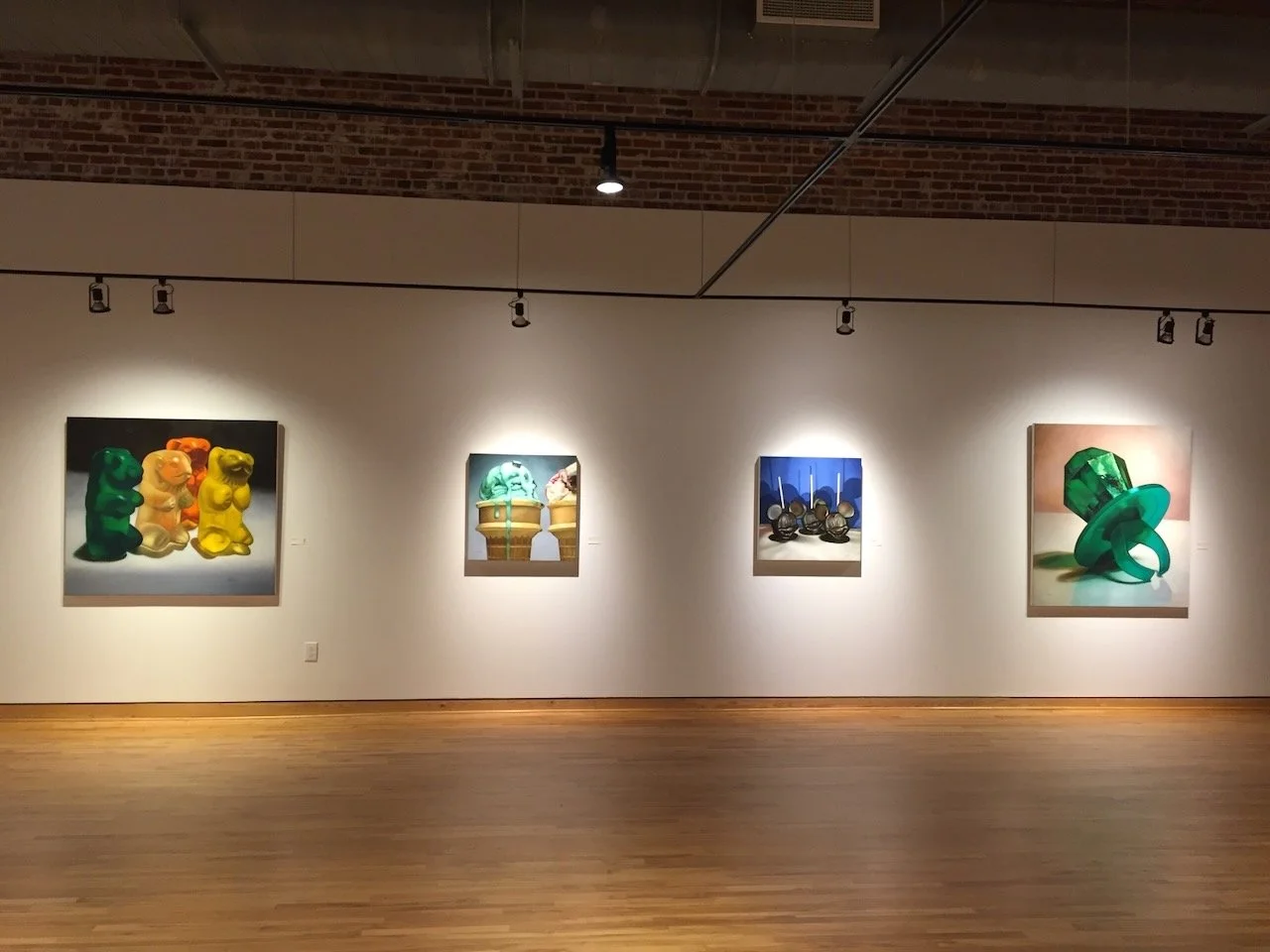 An art gallery with four paintings displayed on a white wall under ceiling-mounted light fixtures. The gallery has a wooden floor and exposed brick ceiling.