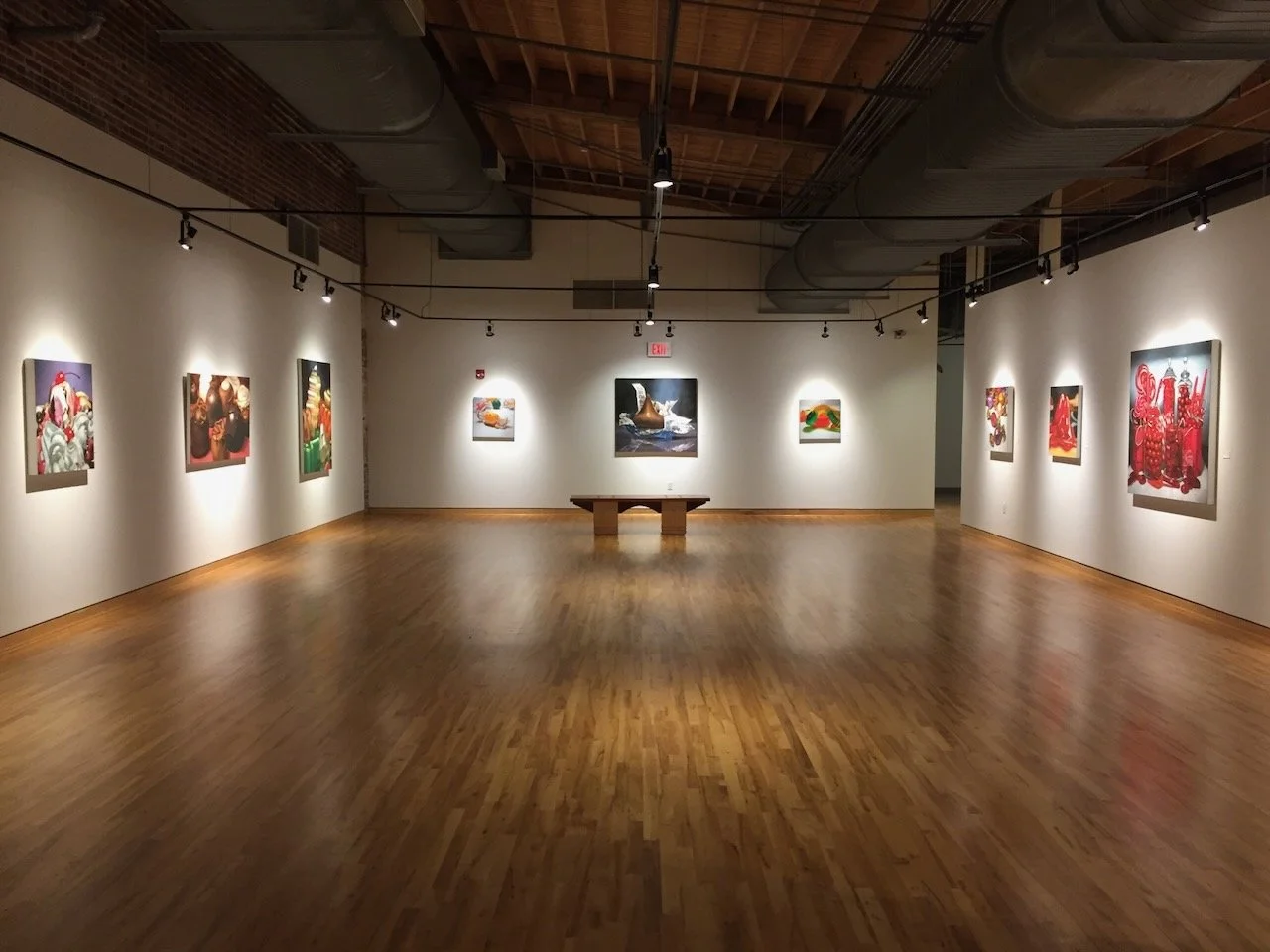 An art gallery with paintings on white walls, wooden flooring, and track lighting. A wooden bench is centered in the room, and the ceiling has exposed ducts and beams.