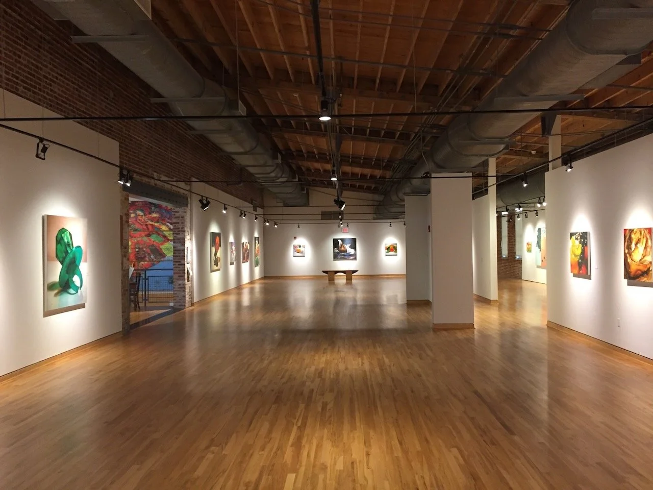 An art gallery with white walls displaying colorful paintings, wooden floors, exposed brick walls, and ceiling ducts, with track lighting illuminating the artworks.