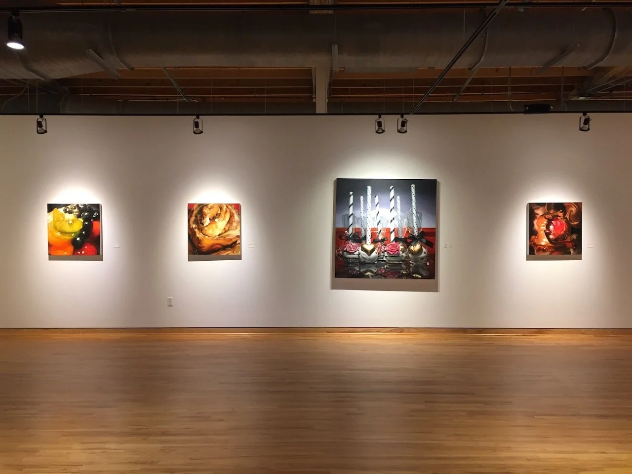 Art gallery wall with five paintings, including fruit and abstract images, illuminated by ceiling spotlights.