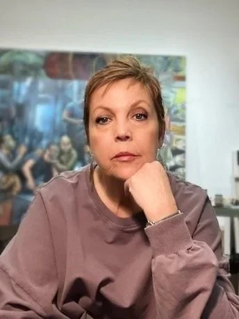 A woman with short hair wearing a mauve sweatshirt, sitting in an indoor space with her hand resting on her chin.