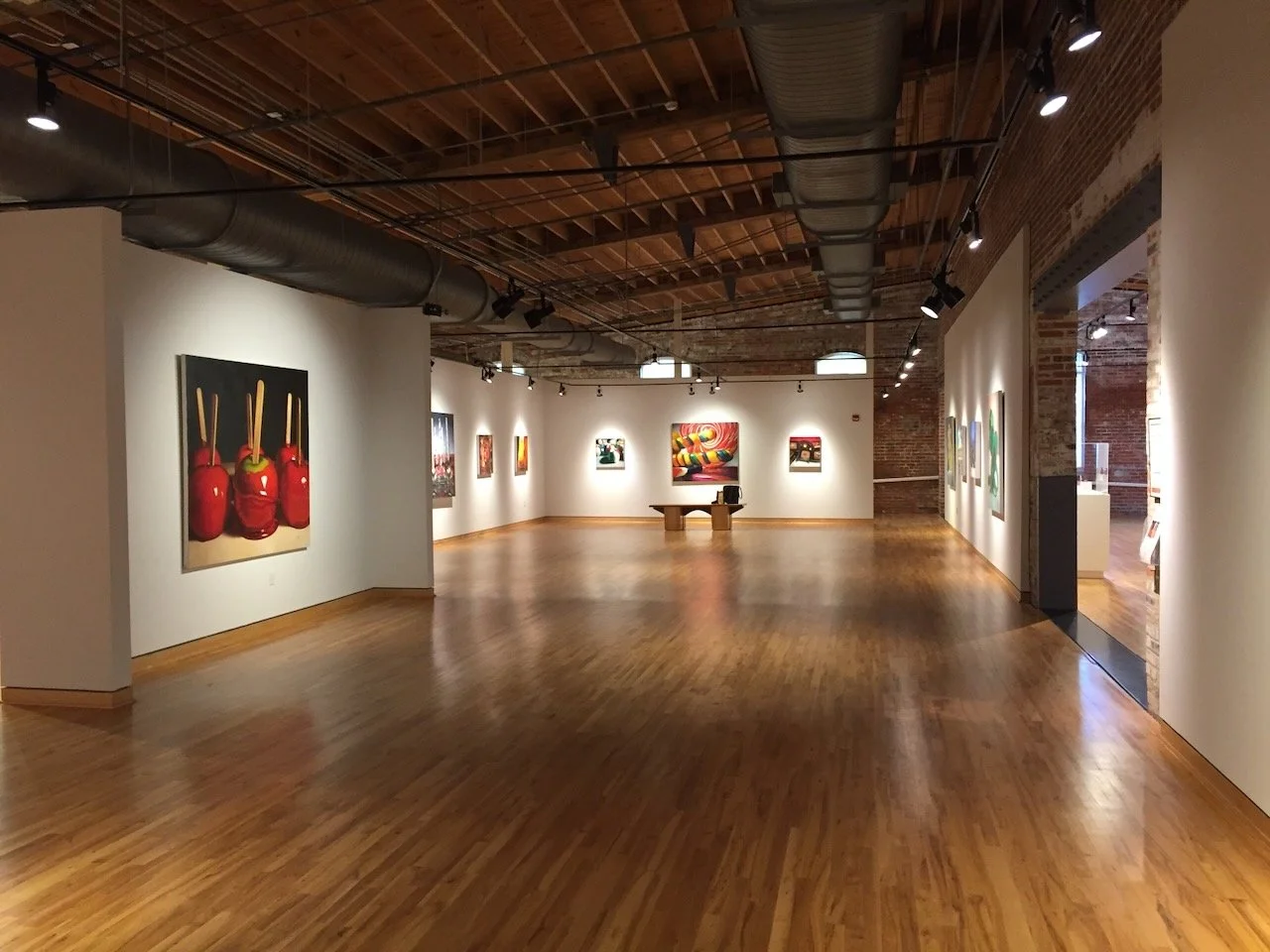 An art gallery with wooden floors and white walls displaying colorful paintings, some abstract and others abstract, with track lighting and exposed brick walls and ceiling.