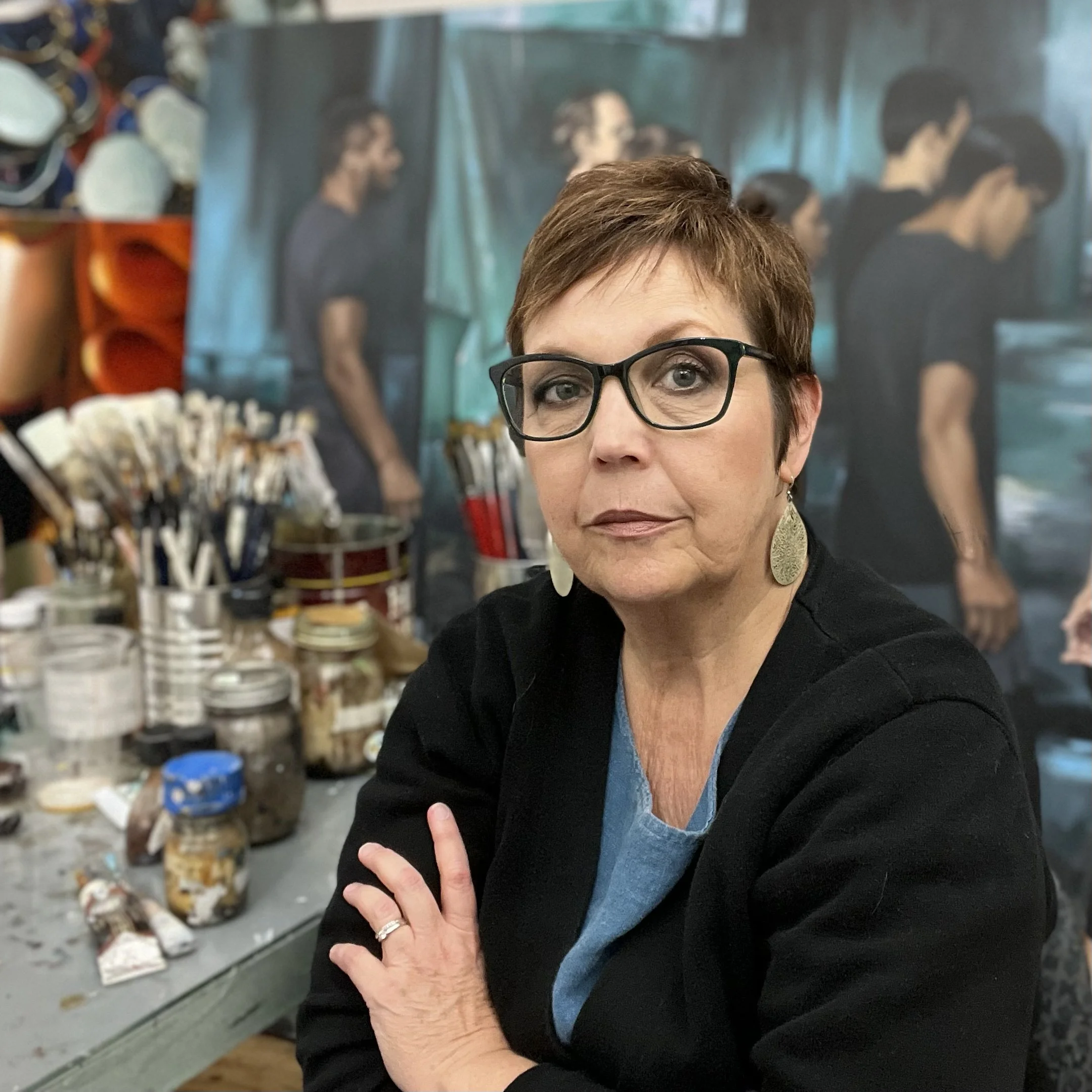 A woman with short brown hair and glasses sitting at a table with art supplies, with a mural of people in black shirts in the background.