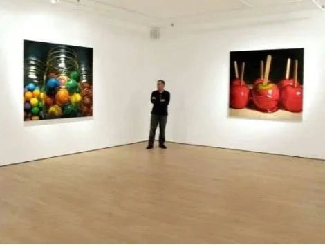 A person standing in an art gallery viewing two large paintings, one of exercise weights and colorful gym balls, and the other of red kettlebells with sticks.