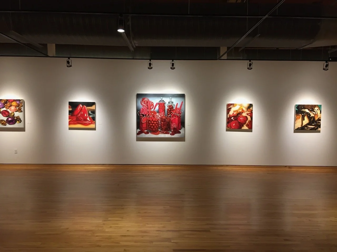 Gallery wall with five colorful still life paintings featuring red and purple tones, illuminated by ceiling spotlights in an art museum with wooden flooring.