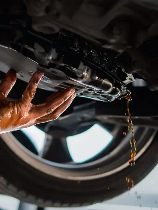 A person inspecting or changing oil in the underside of a vehicle, with oil dripping from the oil pan.