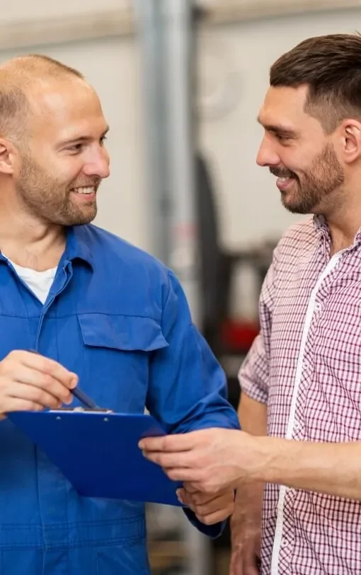 Two men smiling and shaking hands in a workshop, one in a blue uniform holding a clipboard.