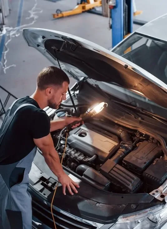 A mechanic inspecting the engine of a car with the hood open in a garage, using a flashlight.