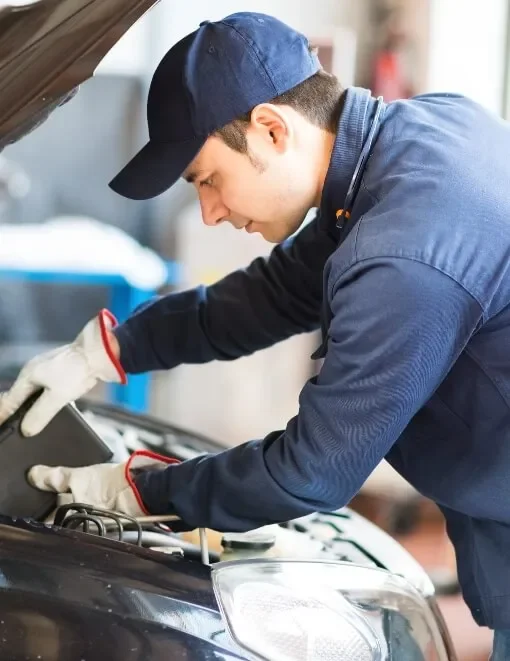A male mechanic wearing a navy blue cap, navy blue work jacket, and white gloves inspecting a car engine in an auto repair shop.