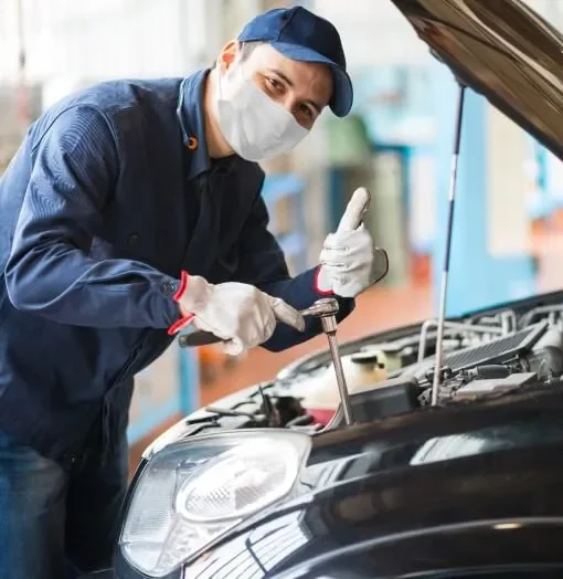 A mechanic wearing a face mask, gloves, and a navy blue uniform, giving a thumbs up while working on a car's engine in a garage facility.