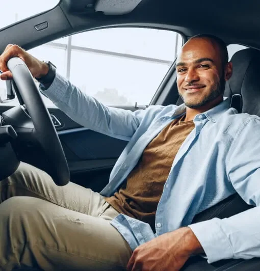 A man sitting in the driver's seat of a car, smiling and wearing a light blue shirt and tan pants.