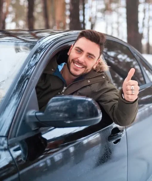 A smiling man in a jacket giving a thumbs-up while sitting in the driver's seat of a car in a snowy forest.