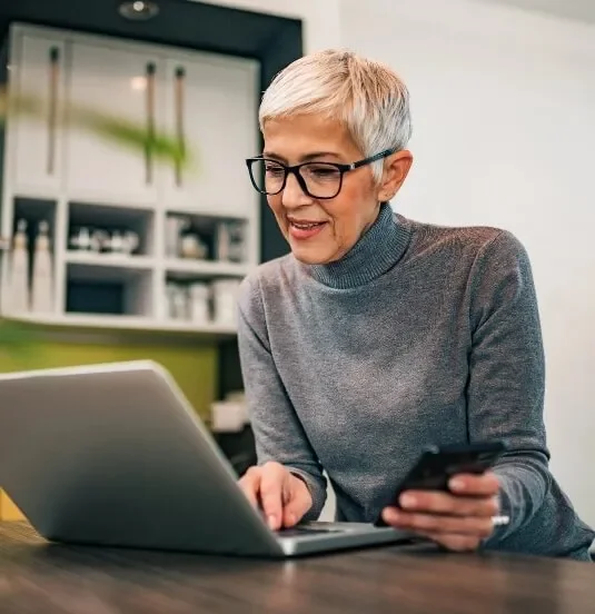 An older woman with short gray hair and glasses using a laptop and holding a smartphone in a modern kitchen.
